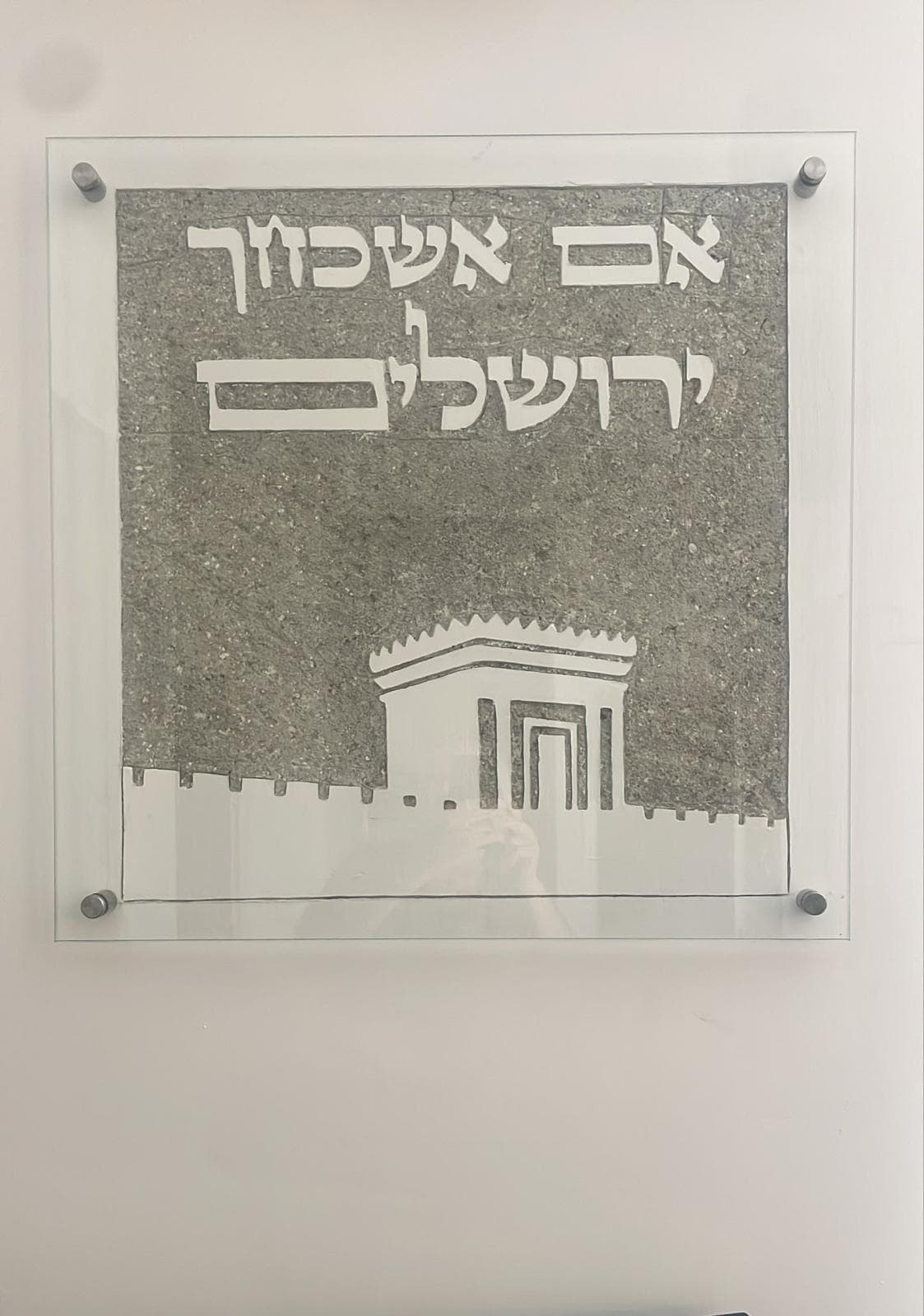 תצוגת גלריה: אריח סיתות מרובע באבן אפורה תלוי על קיר לבן מאחורי לוחית זכוכית שקופה המוחזקת בעמודי מתכת — הכיתוב 'אם אשכחך / ירושלים' חרוט למעלה ובית המקדש על חומות ירושלים מסותת למטה.