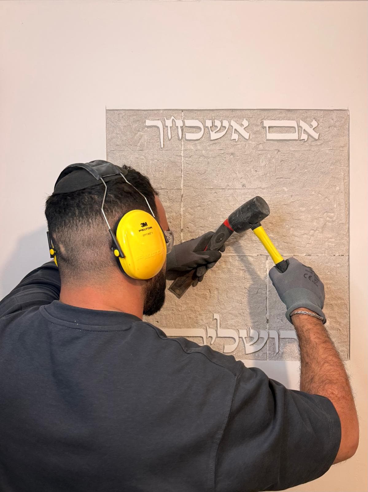 צילום פעולה: גיא, חובש אוזניות הגנה צהובות וכפפות, מקיש בפטיש על אזמל לסיתות קיר במרקם אבן — מעליו מסותת 'אם אשכחך' ומתחת רואים את ראשי האותיות של 'ירושלים' תוך כדי עבודה.