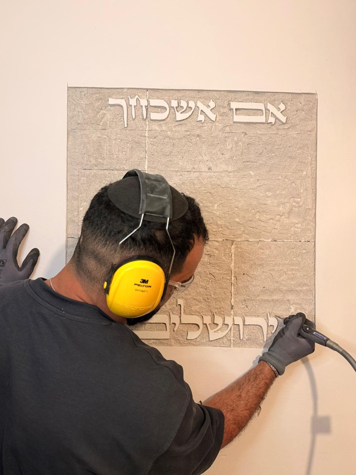 צילום פעולה: גיא, חובש אוזניות הגנה צהובות וכפפות, מסתת בכלי סיבובי את האותיות 'ירושלים' בשורה התחתונה של דגם בקיר — מעליה כבר מסותת בבירור הכיתוב 'אם אשכחך'.