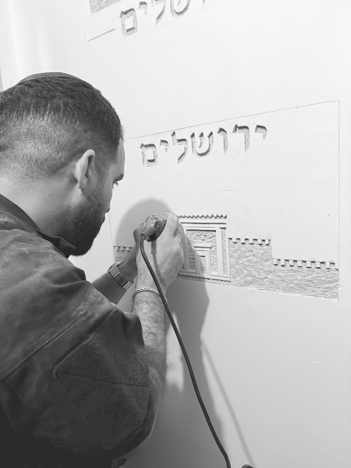 צילום שחור-לבן: גיא בפרופיל, חובש כיפה, מסתת בכלי סיבובי בית מקדש מסוגנן מוקף בחומות ירושלים — מעל אזור העבודה כבר מסותת הכיתוב 'ירושלים' בבירור.