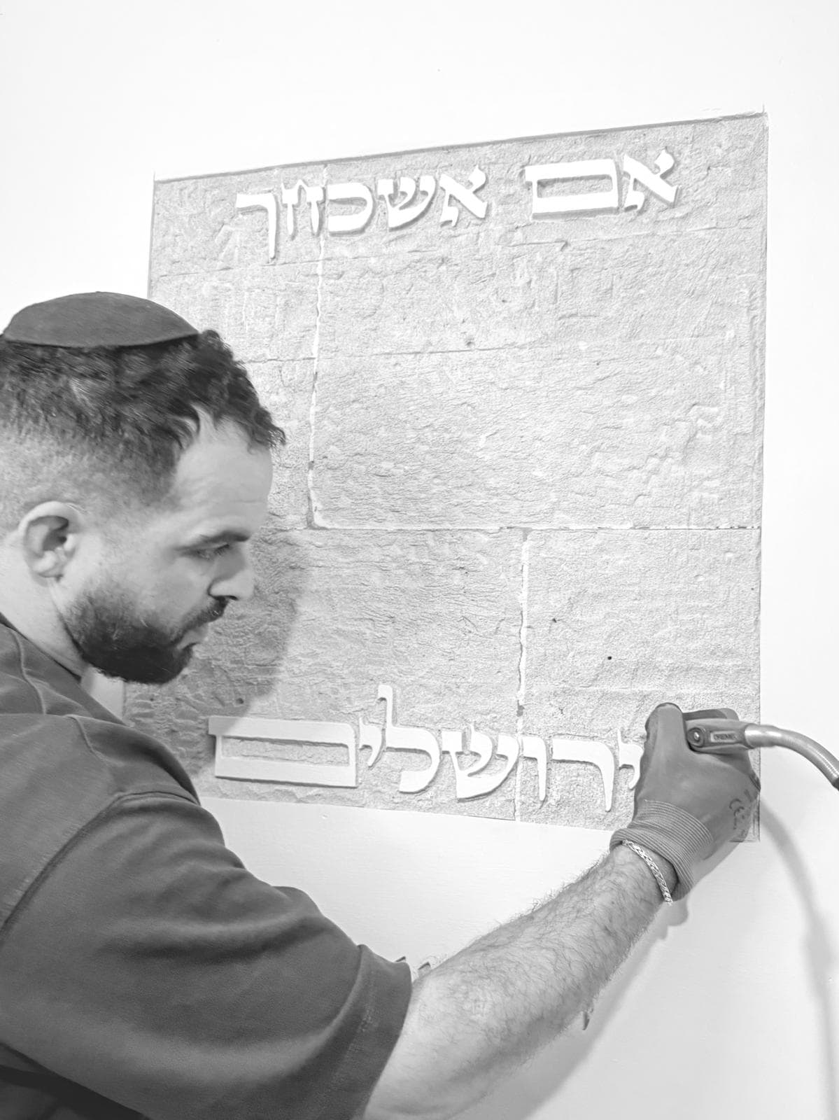 תצלום שחור-לבן: גיא בפרופיל, חובש כיפה, מסתת בידיים עטויות כפפות את האותיות 'אם אשכחך' ו'ירושלים' על קיר חשוף ומחוספס — אחיזה צמודה של הכלי ליד האותיות הגדולות.